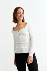 Remera de manga larga, color blanco, con tejido acanalado y escote cuadrado. Presenta un cierre metálico dorado en el centro del escote.