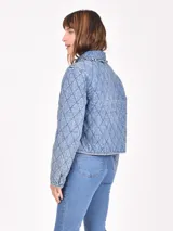 Sobrecamisa acolchada de denim azul claro con patrón de rombos. Presenta cuello con detalle de encaje o puntilla y cierre frontal mediante lazos anudados.
