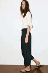 Pantalón de popelina de tiro alto y pernera ancha, de color negro. Presenta pinzas delanteras, bolsillos laterales y falsos bolsillos de vivo en la espalda. Incluye un cinturón combinado tipo pañuelo trenzado con tiras colgantes y cuentas de madera. Cierre frontal con cremallera, botón interior y gancho.