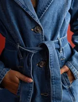 Chaqueta larga de jean azul con cuello con solapa, abertura central con botones, bolsillos delanteros, cintura con pasadores y lazo de ajuste, y puños con botón.