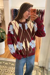 Saco liviano oversize de punto con diseño de rombos argyle y flores celestes sobre fondo marrón, rojo y crema, con cierre frontal de botones y cuello redondo.