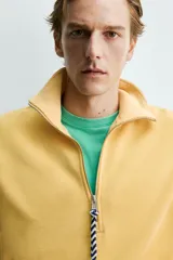 Sudadera de corte relajado confeccionada en algodón con interior de rizo, color amarillo mostaza con efecto lavado. Presenta cuello subido con cierre frontal de cremallera y detalle de cinta trenzada en el tirador, manga larga y acabados en rib.