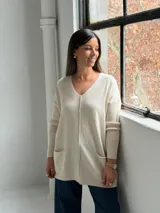 Sweater de punto color beige, con cuello en V, mangas largas con puños acanalados y dos bolsillos frontales. Presenta una costura vertical central y un diseño holgado.