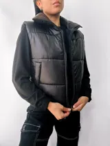 Chaleco puffer negro con cuello alto y cierre frontal con cremallera. Tiene bolsillos laterales y diseño acolchado.