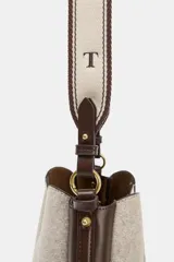 Bolso tipo saca con diseño estructurado, confeccionado en una combinación de tejido de lana en tono beige y laterales de cuero sintético marrón. Presenta costuras decorativas verticales, asa de mano superior y una correa bandolera extraíble con detalle de franjas. Cuenta con cierre mediante botón imantado.