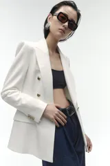 Blazer de corte entallado con cuello y solapa, manga larga y hombros marcados. Presenta un cierre frontal cruzado con botones dorados y bolsillos delanteros con solapa.