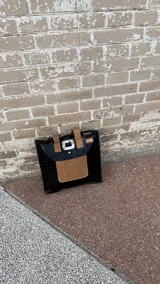 Tote bag de ecocuero negro con textura croco, cuenta con un bolsillo frontal de gamuza marrón y un detalle decorativo circular de guampa en el cierre superior. Posee doble asa de hombro y costuras visibles en los bordes.