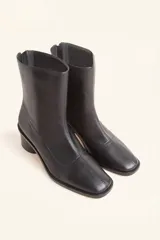 Bota de caña media confeccionada en cuero vacuno negro, con diseño minimalista y cierre trasero. Presenta un taco cilíndrico de madera tallada y punta cuadrada.
