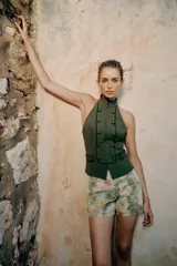 Chaleco verde militar con cuello halter, espalda descubierta y cierre frontal con cremallera oculta. Presenta detalles de falsos botones dorados y ribetes en contraste, con trabillas en los laterales.