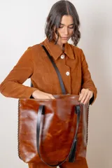 Bolso tipo shopper de cuero vacuno color chocolate con textura efecto croco. Presenta detalles de pasamanería en los laterales, cierre superior y una borla decorativa de flecos de cuero.