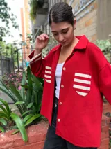 Camisa roja de tejido de algodón con cuello clásico, cierre frontal abotonado, mangas largas con puños acanalados a rayas y dos bolsillos de parche con rayas horizontales en el pecho.