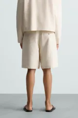 Bermuda de corte relajado color beige claro, confeccionada en tejido con textura. Presenta cierre frontal con cremallera y botón, bolsillos delanteros y bolsillos traseros de doble vivo con botón.