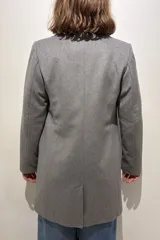 Tapado de paño de lana color gris, con solapas clásicas, cierre frontal de dos botones, bolsillos laterales con solapa y un bolsillo superior en el pecho. Presenta un tajo en la parte posterior y forro interior.