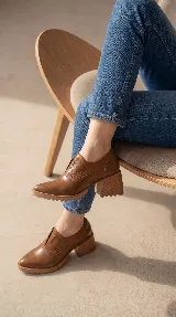 Zapato tipo slip-on color marrón, confeccionado en cuero natural con combinación de texturas lisas y grabadas. Presenta un diseño de punta almendrada, taco cuadrado de madera y suela dentada.