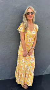 Vestido largo amarillo con estampado floral en tonos rosa, blanco y celeste. Tiene mangas cortas con volados y un volante en el ruedo.