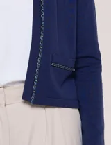 Cardigan corto azul marino de punto, con silueta entallada y largo a la cintura. Cuenta con un ribete decorativo que recorre el cuello redondo y la abertura frontal, integrando una cadena dorada en contraste. Posee bolsillos delanteros simulados y mangas largas.
