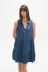 Vestido corto de denim sin mangas, con cuello camisero, cierre frontal con botones y diseño de falda con volados.