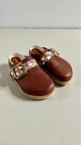 Sandalias de cuero color marrón de la marca Carolina de Cunto, modelo SAKI. Presentan una tira con hebilla metálica grande y detalles pintados a mano en tonos dorados, plateados y blancos. Cuentan con diseño destalonado y suela plana.