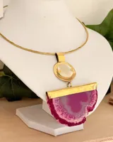 Collar rígido tipo gargantilla realizado en latón dorado con un colgante de gran tamaño que combina una pieza circular de nácar y una piedra ágata en tonos fucsia con terminaciones naturales.