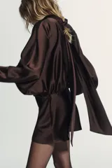 Vestido corto de tejido satinado color marrón chocolate, con cuello subido y lazada tipo foulard integrada. Presenta mangas largas amplias, detalle de frunces en la cintura y cierre lateral con cremallera oculta.