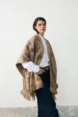 Ruana de tejido de punto con diseño teñido artesanalmente en tonos beige y marrón, con terminación de flecos en el ruedo y etiqueta de cuero sintético en el lateral.