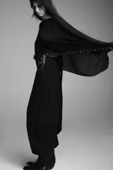 Capa negra de cuello redondo con detalle de tachas plateadas en el ruedo y pañuelo tipo bufanda integrado en el mismo tejido.