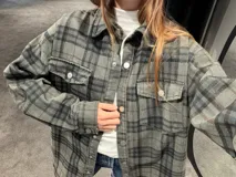 Camisa tipo sobrecamisa de corte oversize, confeccionada en tela a cuadros en tonos marrones y negros. Presenta cierre frontal con botones metálicos a presión, bolsillos frontales con solapa y mangas largas con puños abotonados.