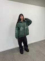 Buzo con capucha y cierre frontal completo, color verde oscuro con efecto lavado degradado. Presenta estampado gráfico en tipografía estilizada en el frente y mangas, bolsillos laterales y etiqueta de marca en el puño.