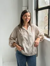 Camisa de lino color beige con diseño oversize, cuello camisero mao, cierre frontal con botones ocultos, mangas largas abullonadas con puños elásticos y delicados bordados florales tono sobre tono en el frente y las mangas.