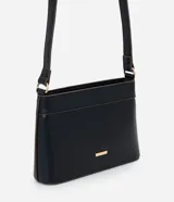 Bolsa bandolera pequeña de estructura firme, confeccionada en poliuretano color negro con detalles de costuras en contraste. Cuenta con cierre metálico, asa larga ajustable con hebilla y placa metálica decorativa con el logo de la marca.