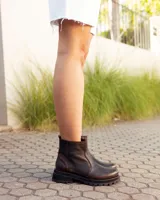 Bota de caña alta confeccionada en cuero color marrón caoba con acabado desgastado. Presenta una suela track robusta, correas con hebillas en la parte superior de la caña y un arnés decorativo con herrajes metálicos en el tobillo.