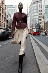 Chaqueta color beige con efecto ante, cuello redondo, manga larga y cierre frontal con cremallera. Presenta un bajo en evasé y un cinturón fino de cuero marrón oscuro con hebilla metálica.