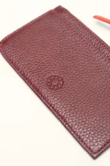 Monedero rectangular de cuero vacuno con textura granulada en color marrón topo. Cuenta con cierre superior metálico y un tirador decorativo de cuero en color naranja con un nudo. Presenta un pequeño logo grabado en el centro.
