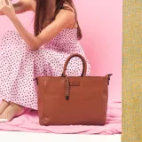 Cartera de formato amplio y estructurado, color marrón, con doble asa de mano y correa larga desmontable y regulable que permite llevarla al hombro o cruzada. Cuenta con cierre superior y aplique decorativo en el frente.
