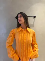 Camisa de manga larga color naranja, con cuello clásico y cierre frontal de botones. Presenta detalles decorativos verticales con textura en el frente y en los bordes del cuello, además de mangas con volumen y puños ajustados.