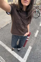 Camiseta de algodón color marrón oscuro, de corte regular, cuello redondo y manga corta. Presenta un detalle de lazos con tejido en relieve en el frente.