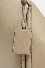 Bolso tipo shopper de hombro en color beige, con diseño estructurado, doble asa y detalle de pespuntes verticales en el cuerpo. Incluye un colgante decorativo tipo etiqueta y cierre imantado.