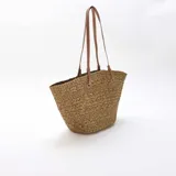 Bolso tipo capazo color kaki, confeccionado en tejido trenzado efecto rafia, con doble asa larga de hombro en cuero sintético marrón claro con costuras blancas visibles.