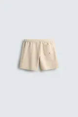 Bañador largo de hombre color rosa pálido con textura seersucker. Presenta cintura elástica ajustable con cordón blanco, bolsillos laterales y un bolsillo trasero de plastrón.