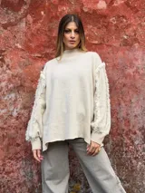 Sweater de punto color beige, con cuello alto, mangas largas abullonadas y detalle de flecos en las costuras de las mangas.
