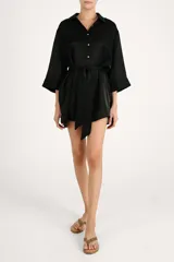 Camisa larga de satén negro, fluida, con cuello camisero, cierre frontal de botones dorados, mangas tres cuartos y lazo ajustable en la cintura.