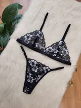 Conjunto de lencería compuesto por un corpiño triangular y una bombacha tipo tanga, confeccionados en encaje negro con detalles florales en hilo plateado.