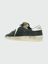 Zapatillas Golden Goose modelo Stardan, confeccionadas en gamuza verde oscuro con detalles en cuero liso azul marino en la estrella lateral. Presentan cordones blancos y una suela de goma desgastada color crema. El lateral lleva la inscripción manuscrita "Golden Goose" y la referencia "GGDB/STARDAN".