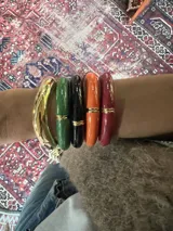 Set de pulseras rígidas de diferentes colores y materiales, incluyendo una pulsera dorada tipo brazalete y pulseras de resina en verde, negro, naranja y borgoña, con detalles dorados.