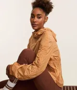 Campera deportiva femenina tipo parka, color ocre, confeccionada en poliamida y poliéster. Tiene cuello con capucha, mangas largas con puños elásticos, cierre frontal con cremallera y cordón ajustable en la cintura.