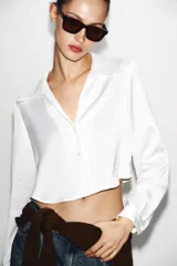 Camisa corta de tejido satinado color marrón, con cuello solapa, escote en pico y manga larga. Presenta cierre frontal con botones forrados a tono.