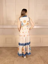 Vestido largo color crema con estampado de flores y corazones en tonos azules, naranjas y amarillos. Tiene cuello en V con ribete azul y corte imperio con falda escalonada.