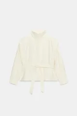 Blazer corto de color marfil con cuello subido, manga larga y bolsillos laterales. Cuenta con un cinturón del mismo tejido para anudar en la cintura, forro interior a tono y cierre frontal con cremallera y botones a presión ocultos bajo solapa.
