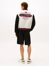 Campera estilo universitario de Tommy Hilfiger, colección APXGP, color blanco con mangas negras y detalles en rojo. Confeccionada en satén aterciopelado, con cuello estilo béisbol, cierre a presión, bolsillos laterales ribeteados, cuello, puños y bajo de punto canalé con rayas, logo de la colección APXGP bordado en la parte delantera y parches con el logo de la colección APXGP en mangas.