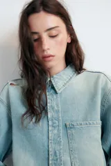 Cazadora corta de denim en tono celeste claro, con cuello solapa, manga larga y cierre frontal con botones. Presenta un bolsillo de plastrón en el delantero y detalles de rotos en el cuello y el ruedo deshilachado.